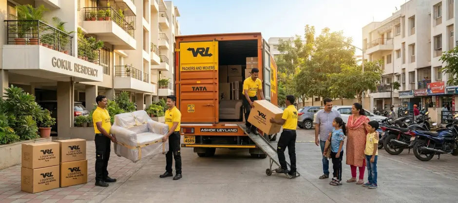 Panoramic view of household relocation outside an Indian apartment building. An orange-and-yellow Ashok Leyland container truck with VRL Logistics Packers an…